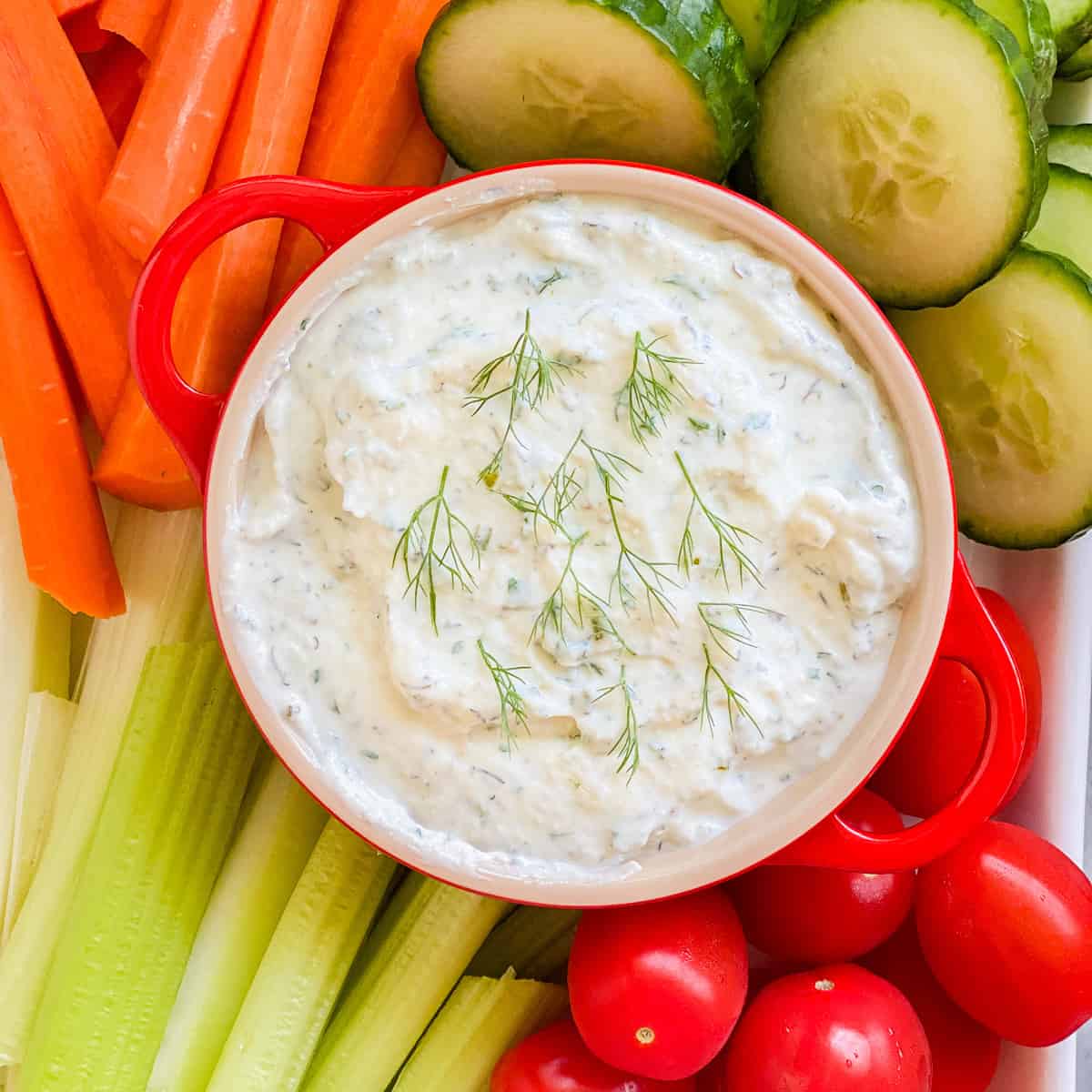 Red crock with white dip surrounded by veggies.