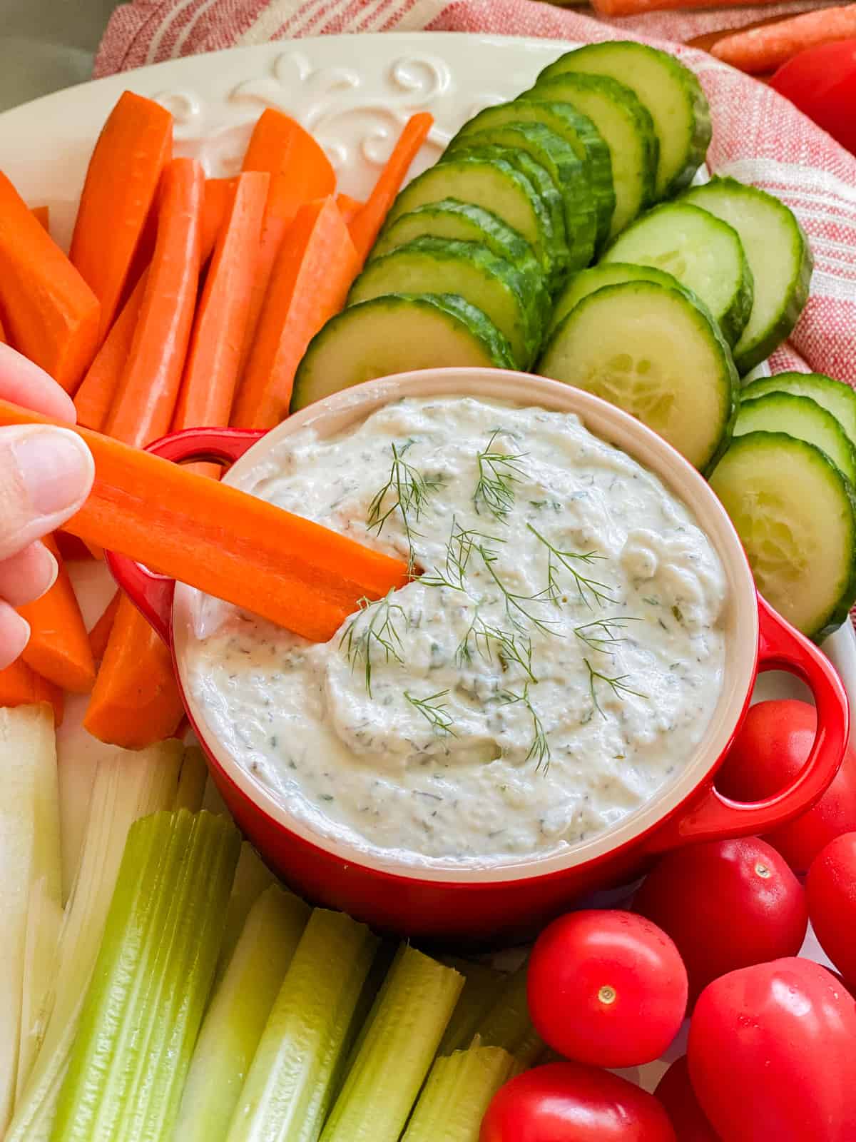 Female hand holding a carrrot stick dipping into a red crock filled with white dip.