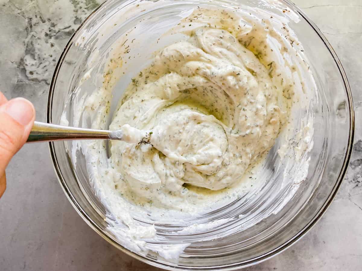 Glass bowl filled with white dip and a hand stirring with a spoon.