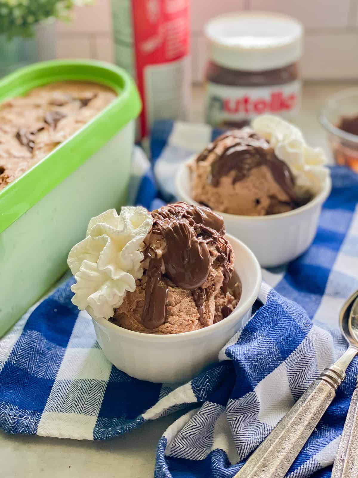 Two white ramekins with chocolaate ice cream, chocolate topping, and whipped cream on a blue checkered cloth with spoons.