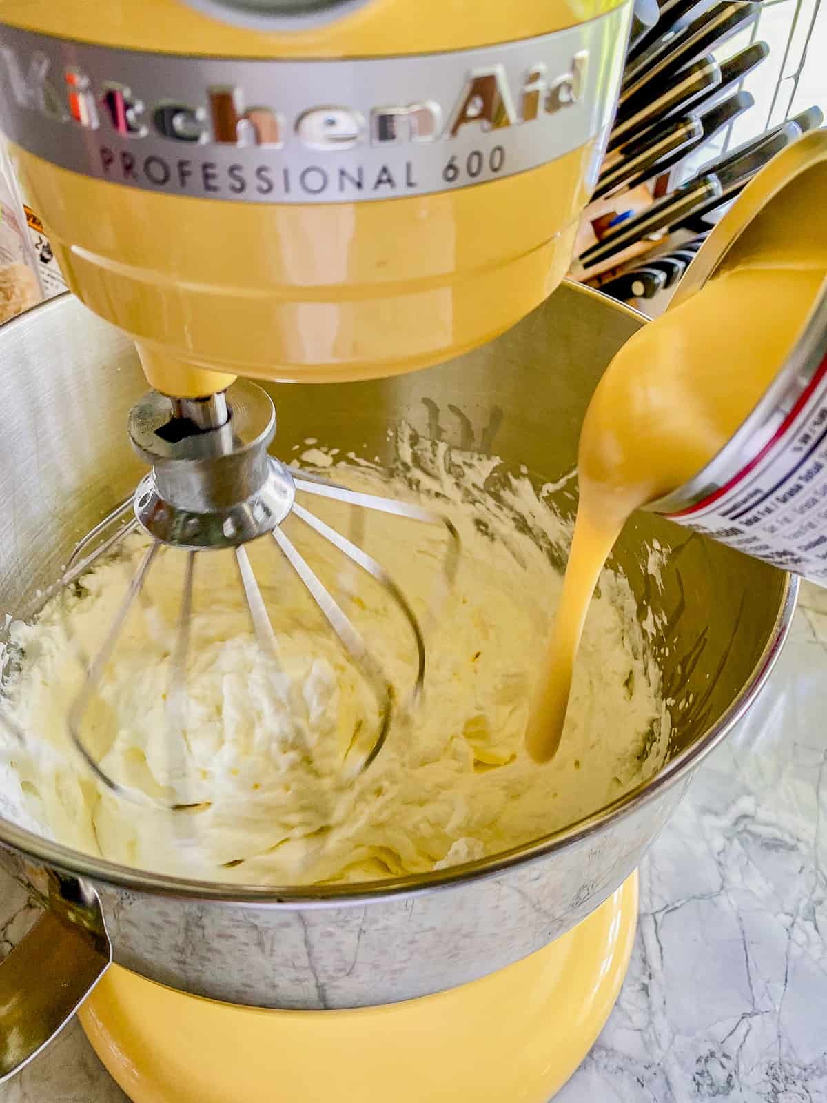 Yellow KitchenAid stand mixer with a balloon whisk whipping cream and pouring in condensed milk.