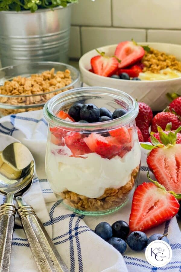 Glass jar filled with a layer of granola, yogurt, and fruit with fruit and spoons on the side.
