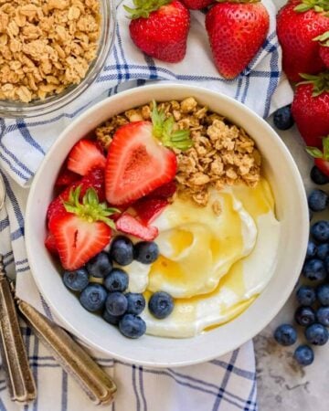 White bowl filled with yogurt, honey, bleuberries, strawberries, and granola with spoons and a striped white and blue napkin.