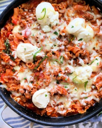 Black saute pan with lasagana cooked inside resting on a striped cloth.