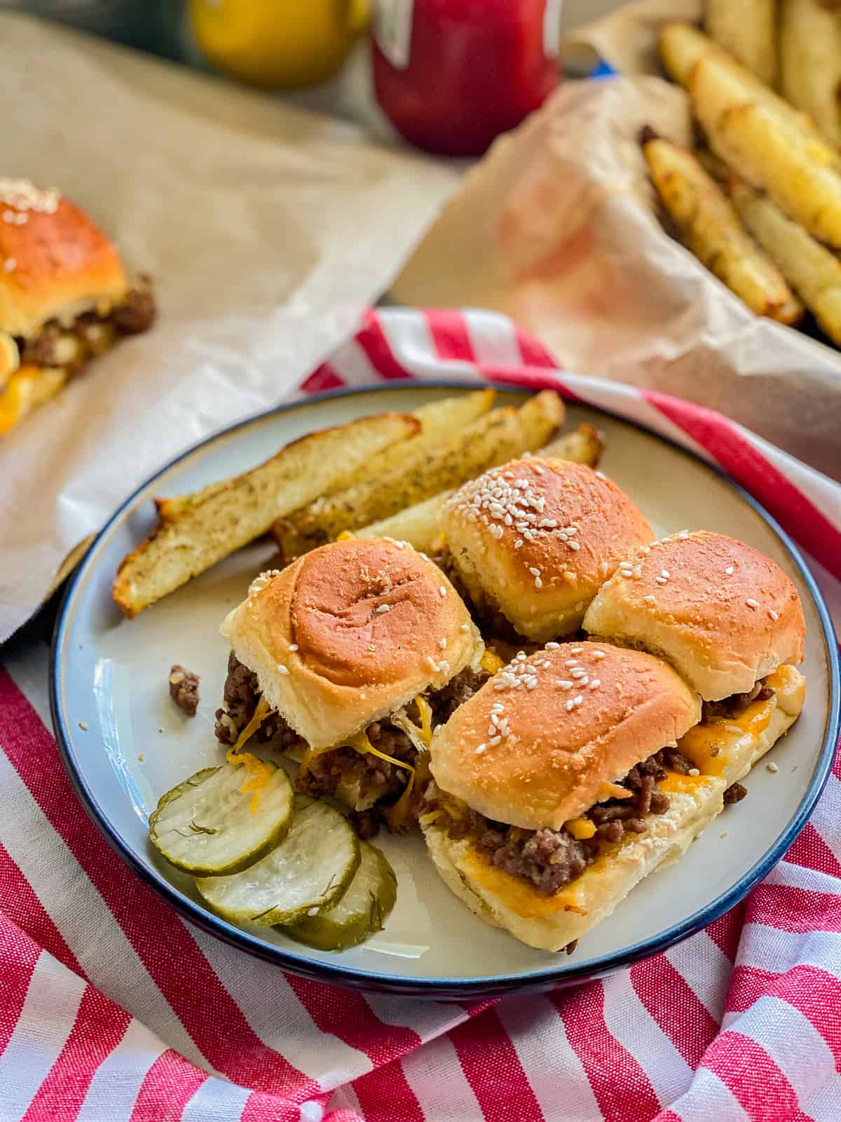 White plate filled with 4 sliders with 3 pickle slices and potato wedges on a red and white striped cloth.