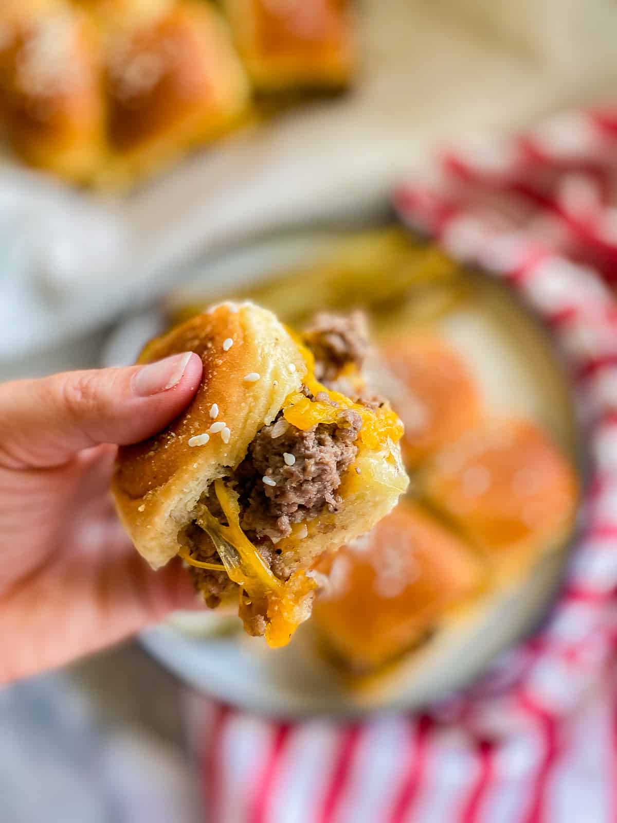Hand holding a ground beef slider with cheese over a plate of sliders.