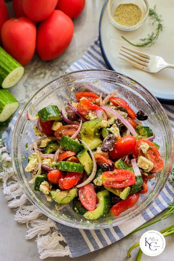 Glass bowl filled iwth cucumber tomato salad on a blue and white stripe cloth with logo on right corner.