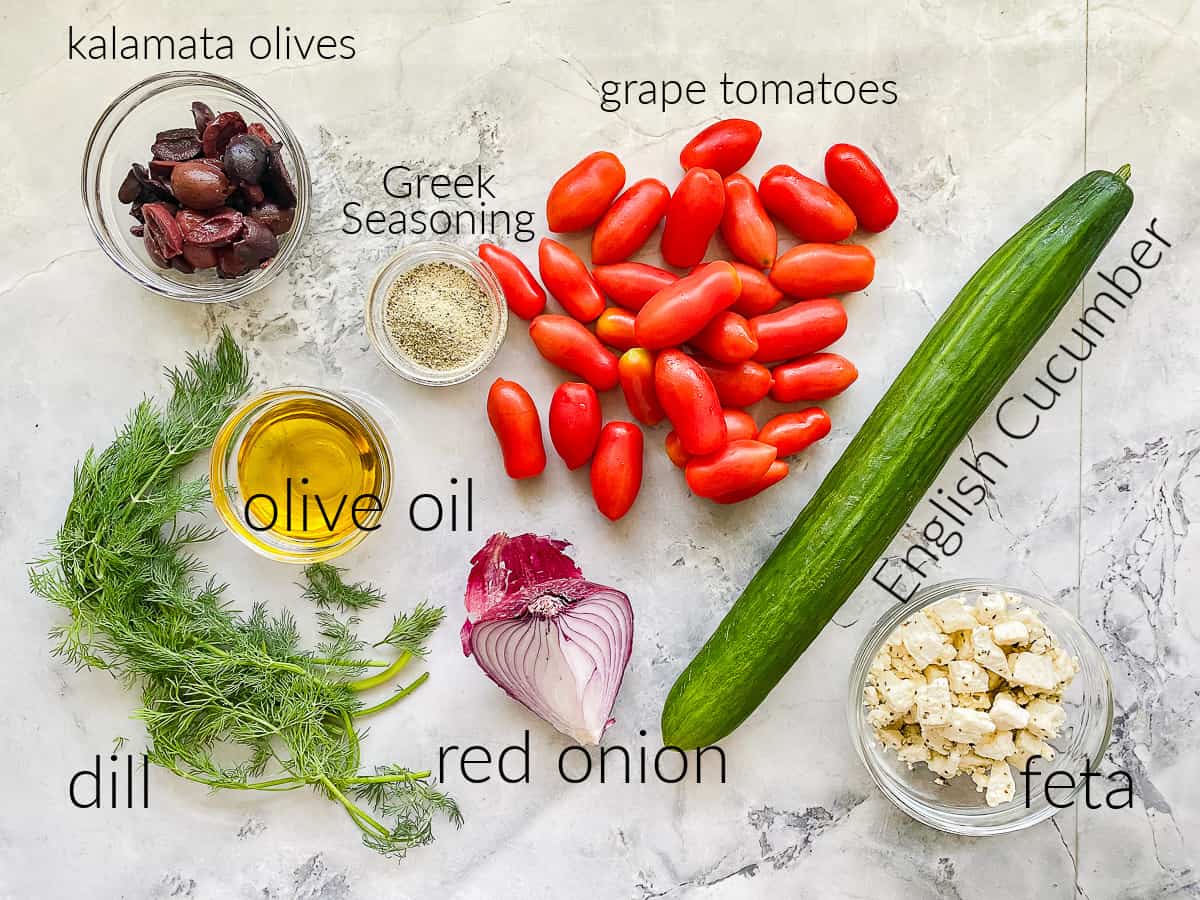 Ingredients on marble counter: olives, dill, oil, Greek Seasoning, cucumber, tomato, red onion, and feta.