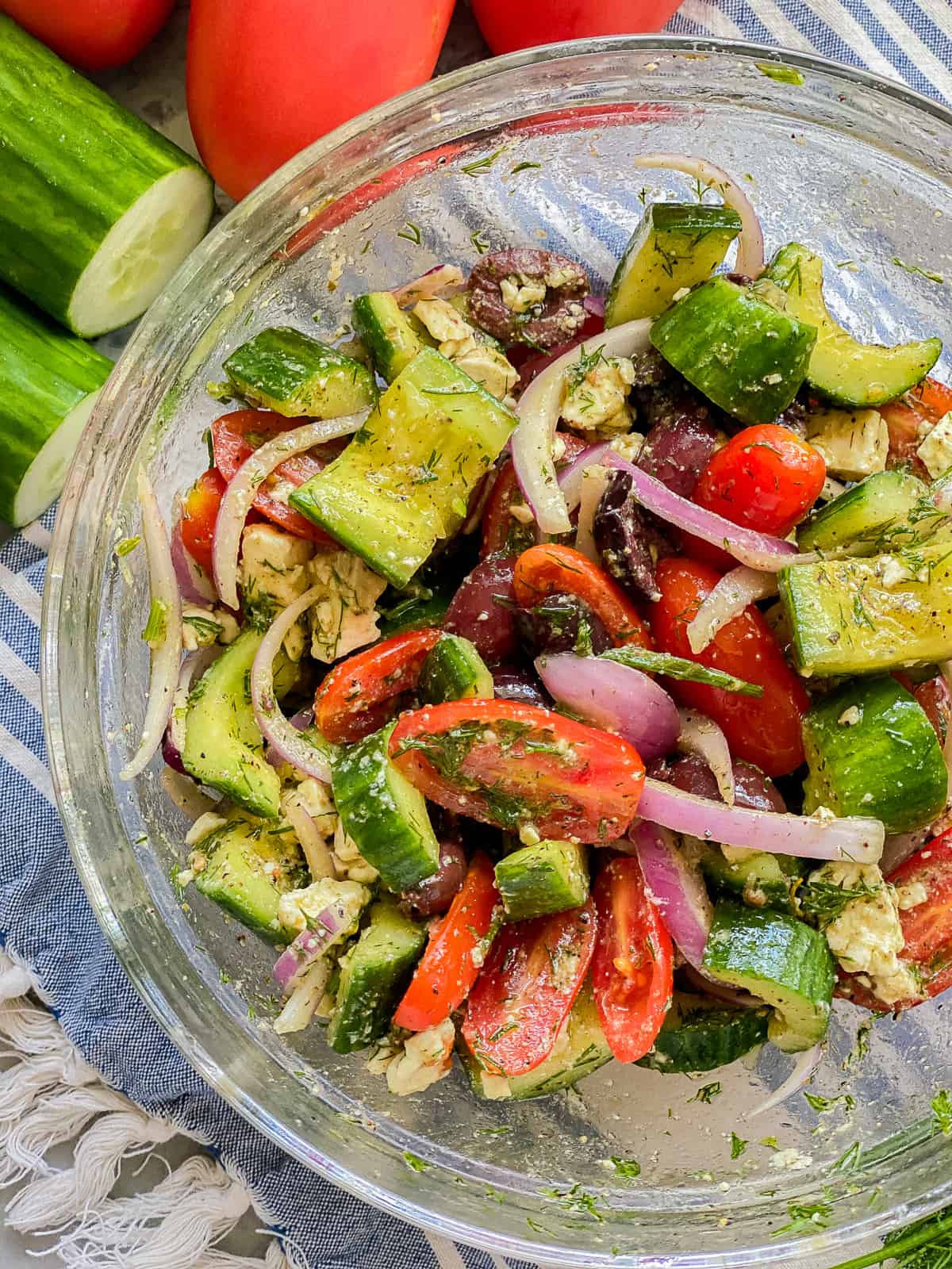 Glass bowl filled with cucumber, tomato, onion, and feta on blue and white striped cloth.