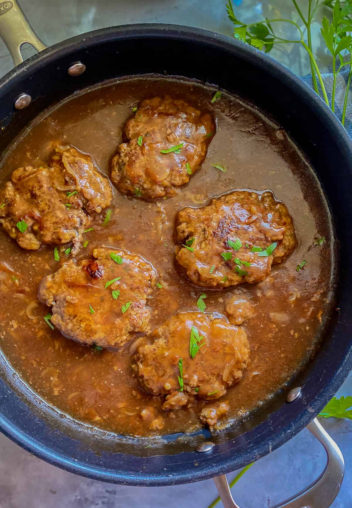 Black skillet with five beef patties in gravy with parsley sprinkled on top.