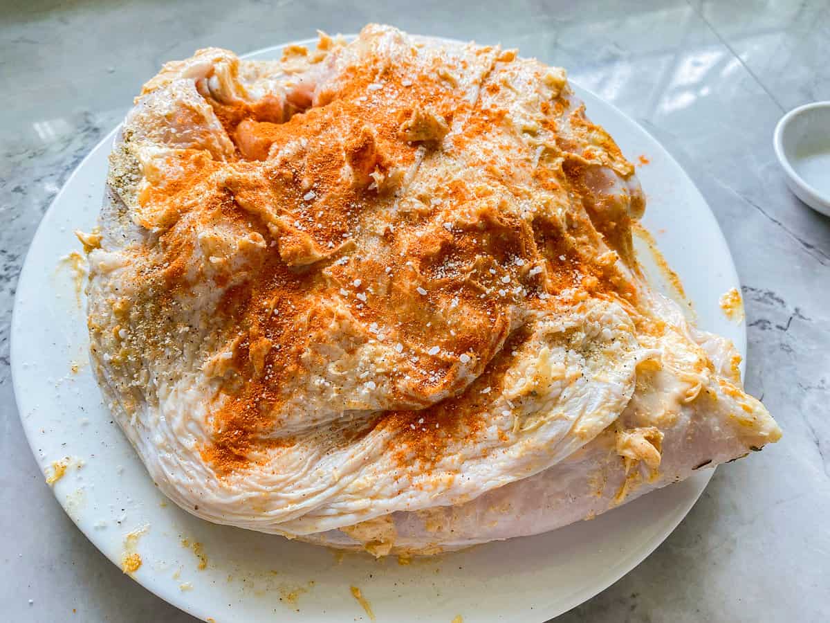 Raw turkey breast on a white plate with butter and seasonings smeared on top.