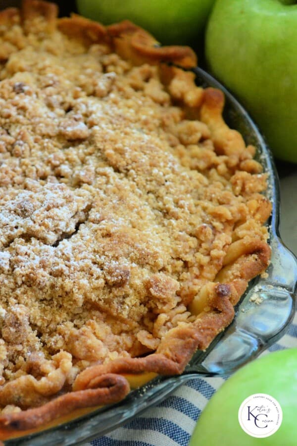 Glass pie plate filled with a pie with a crumb topping and green apples in the background with logo on right corner.