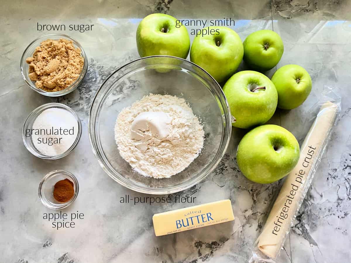 Ingredients on counter: sugar, apple pie spice, flour, apples, butter, and pie crust.