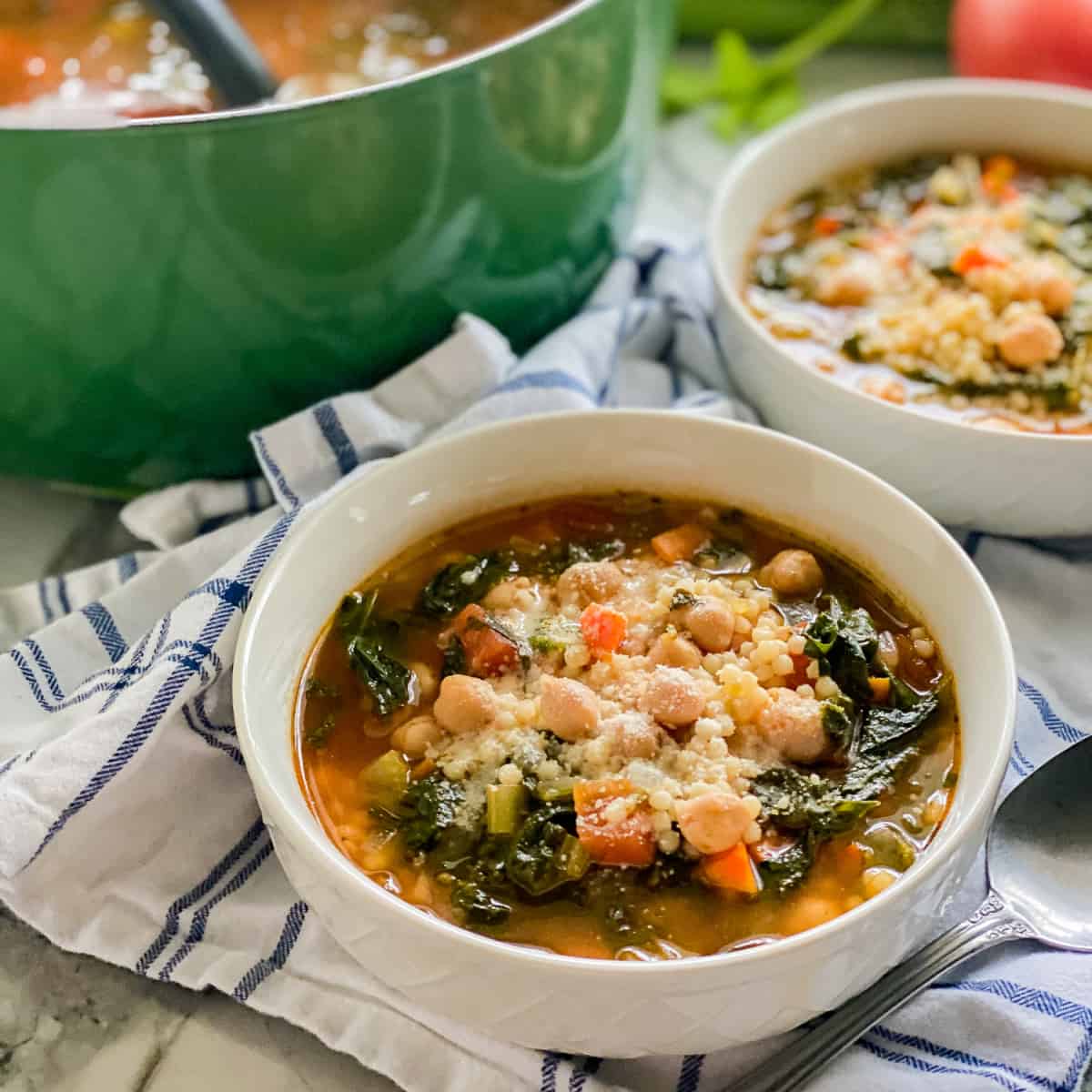 Two white bowls filled with veggies soup with chickpeas and a green pot in the background.