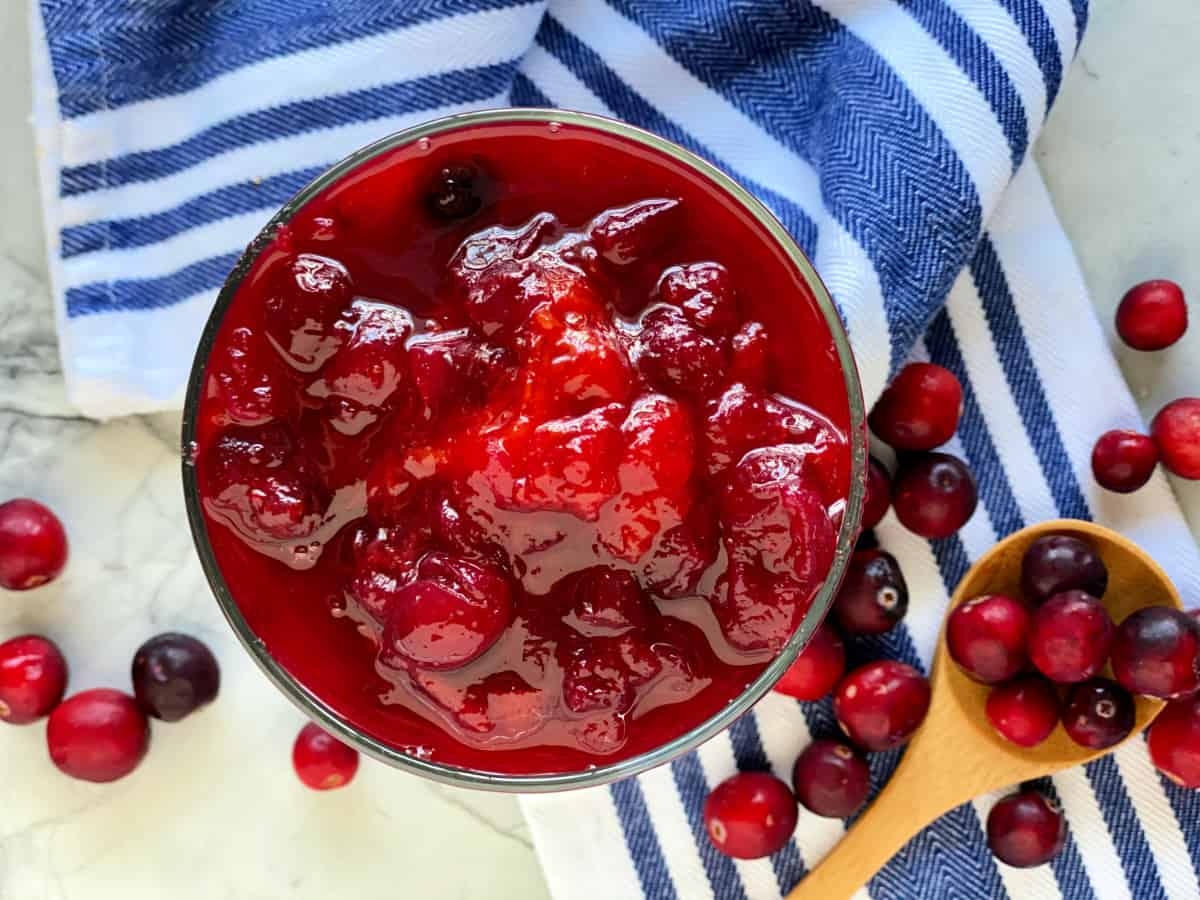 Glass bowl with mango cranberry sauce with a wooden spoon next to it with fresh cranberries.