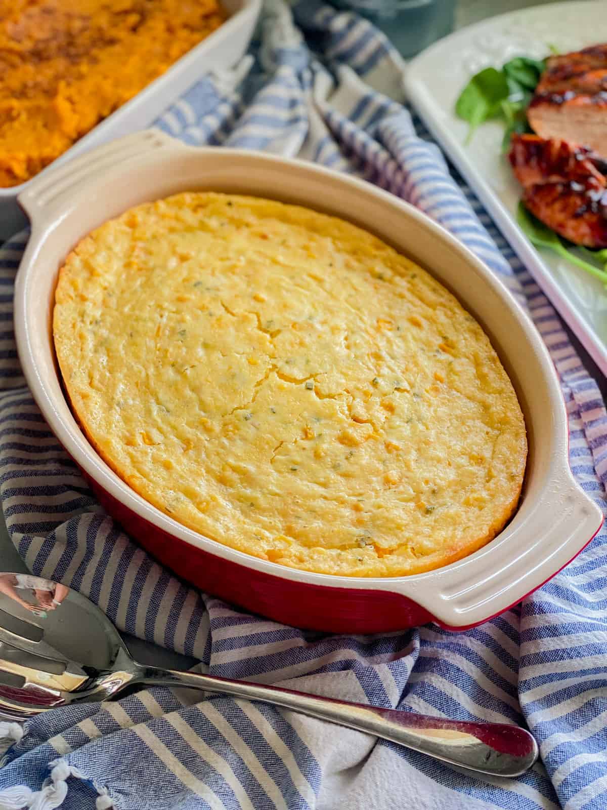 Red oval baking dish filled with corn cassserole resting on a blue and white striped towel.