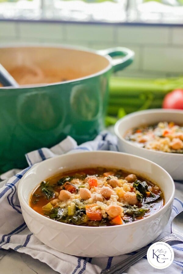 Two white bowls filled with veggie soup and chickepeas with a green pot in the background with a logo on the right corner.