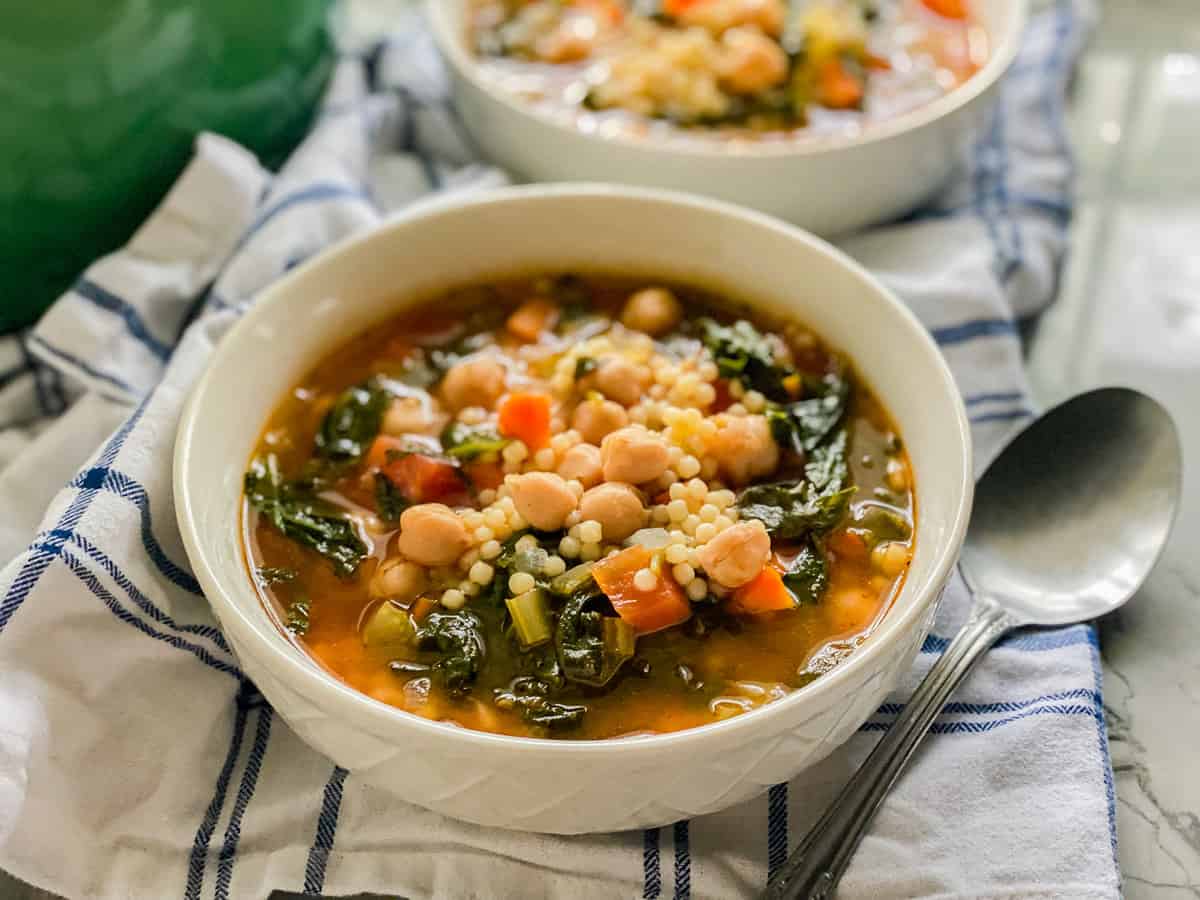 Two white bowls filled with vegetables, couscous, and broth with a spoon on the side.