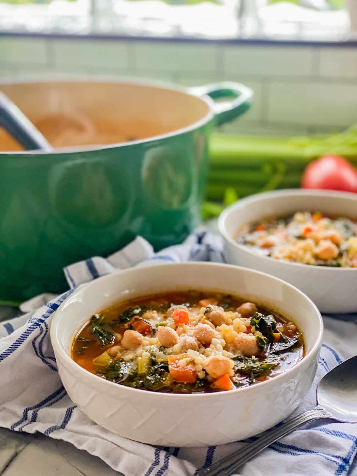 Two white bowls filled with vegetable soup with a green pot.