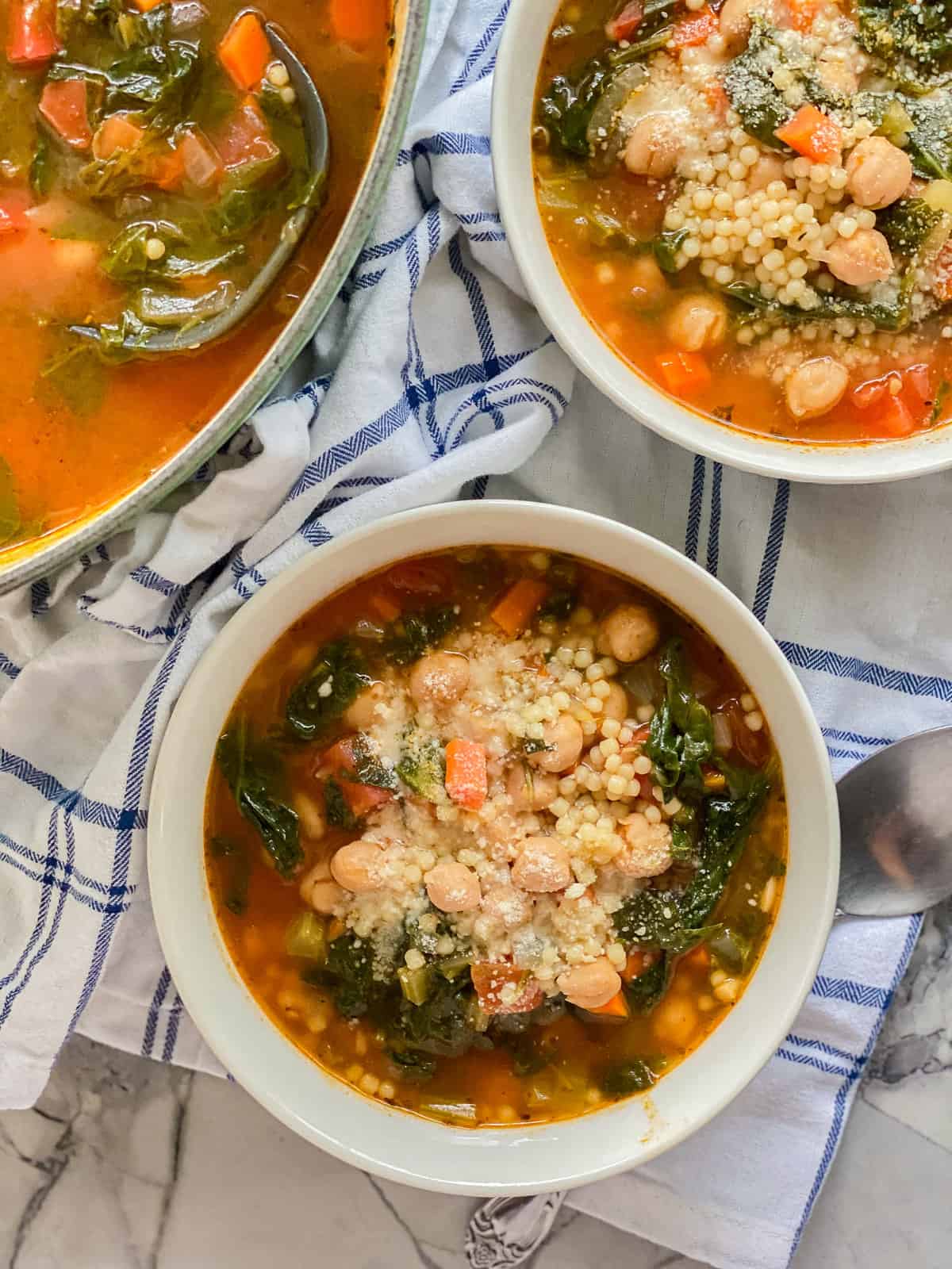 Two white bowls with veggie soup and chickpeas with a pot next to it.