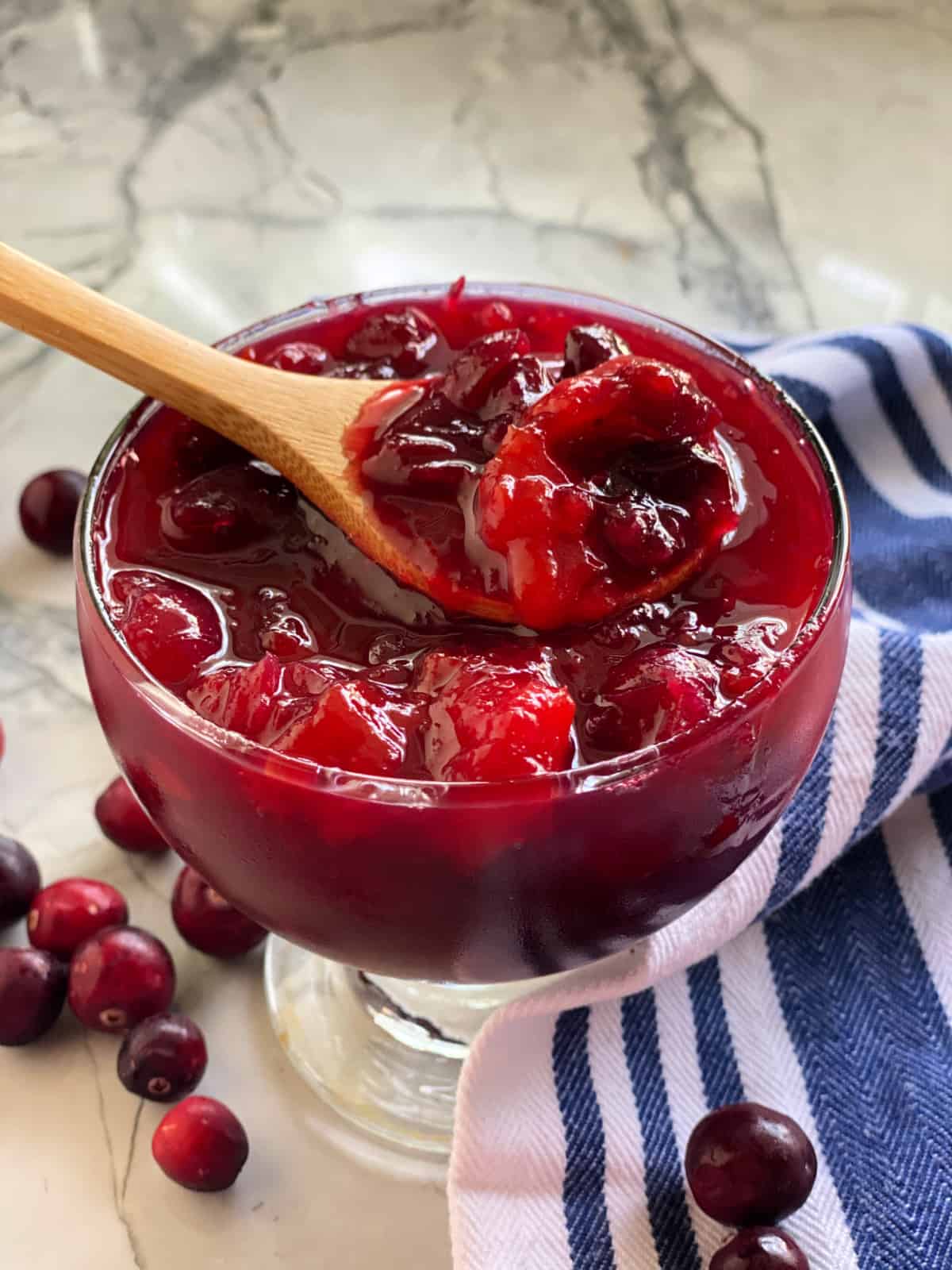 Glass bowl filled with wooden spoon with mango cranberry sauce on a marble countertop.