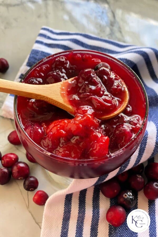Glass bowl filled with cranberry sauce with a wooden spoon dipping into the bowl with logo on right corner.