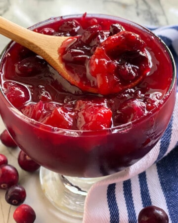 Glass bowl filled with wooden spoon with mango cranberry sauce on a marble countertop.