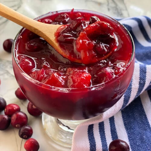 Glass bowl filled with wooden spoon with mango cranberry sauce on a marble countertop.