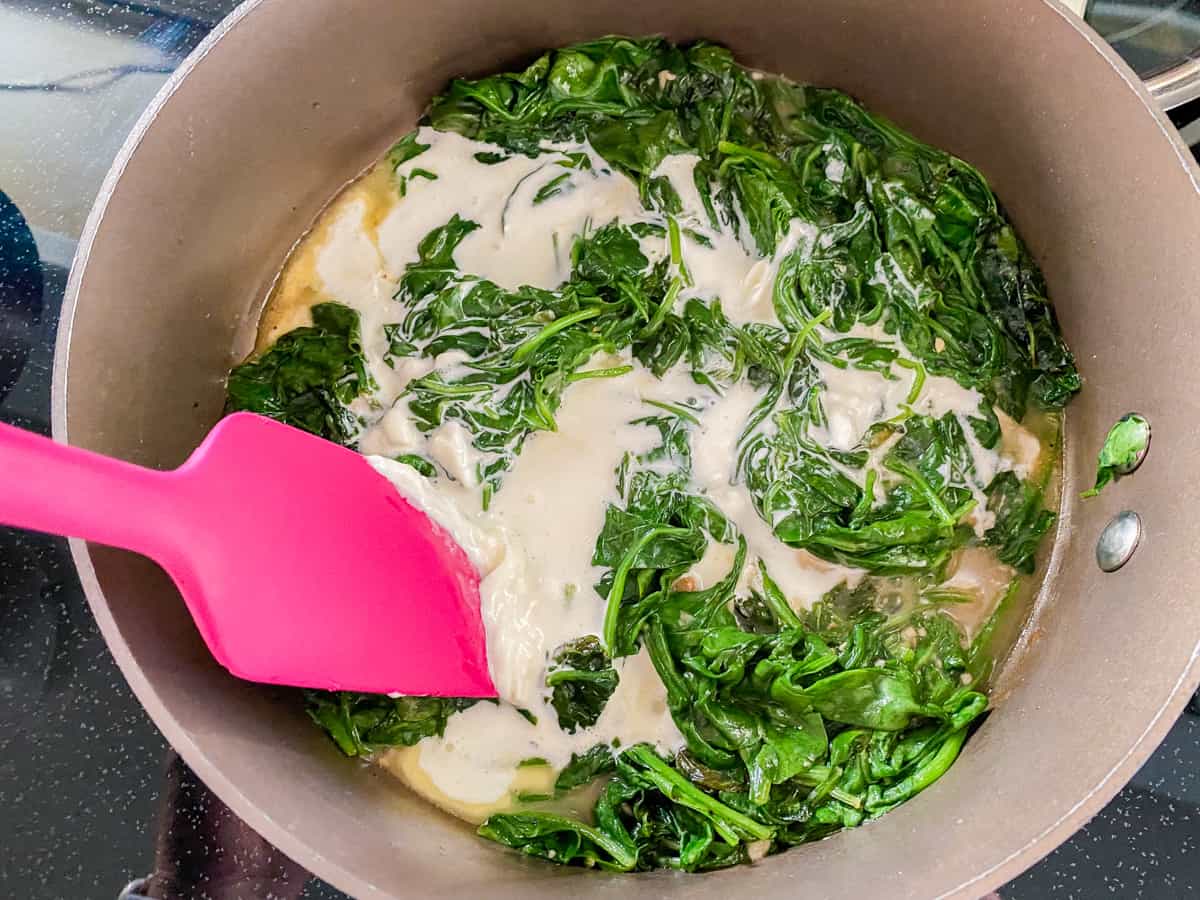 Brown pot filled with wilted spinach, cream, and pink spatula.
