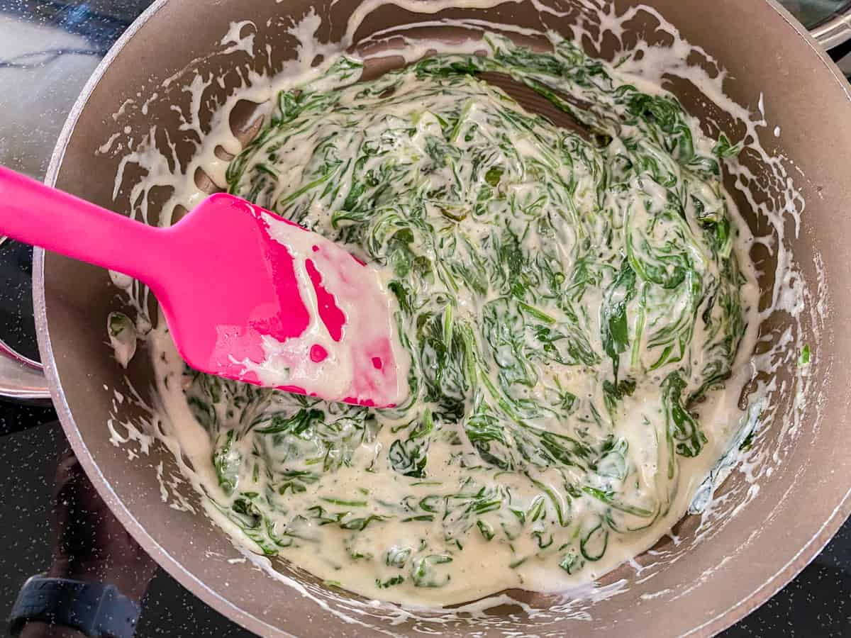 Brown pot filled with creamed spinach with a pink spatula.
