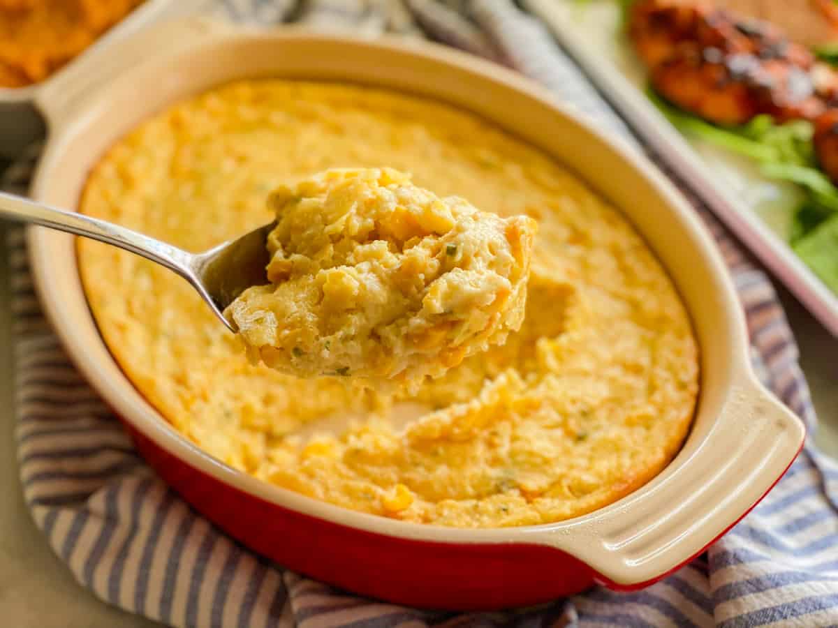 Red oval baking dish with a spoon holding corn casserole.