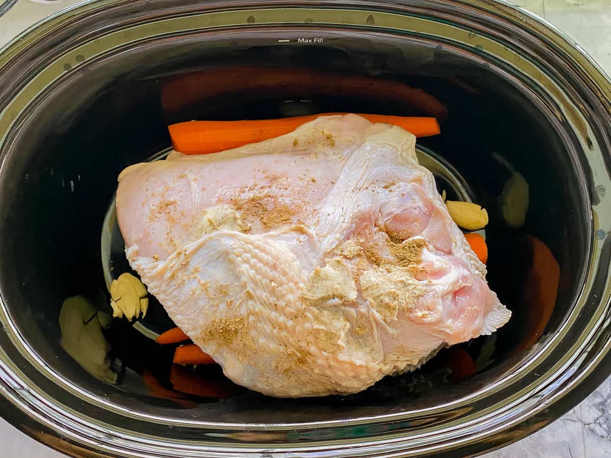 Black oval slow cooker with a raw turkey breast with carrots and garlic in it.