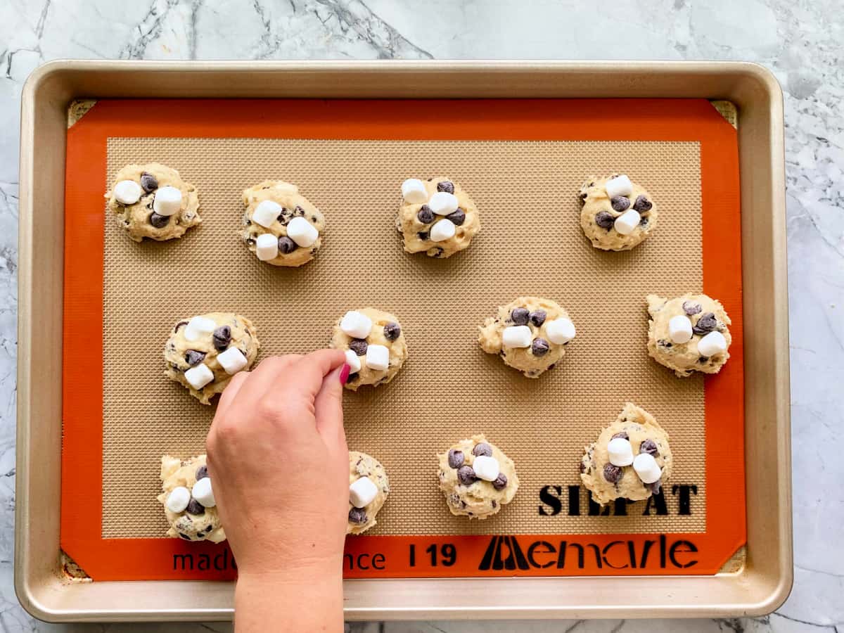 Hand pushing marshmallows onto raw cookie dough balls on a golden baking sheet.