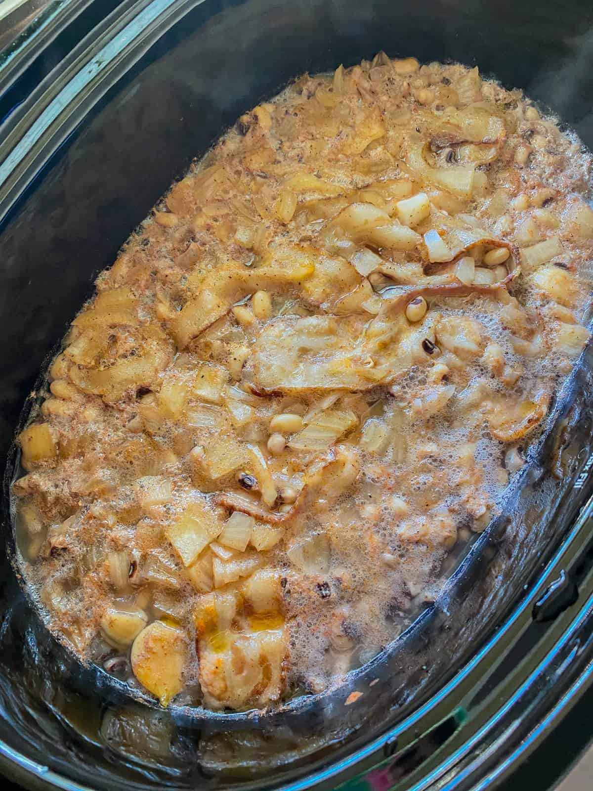 Black oval slow cooker with cooked onions, garlic, and beans.