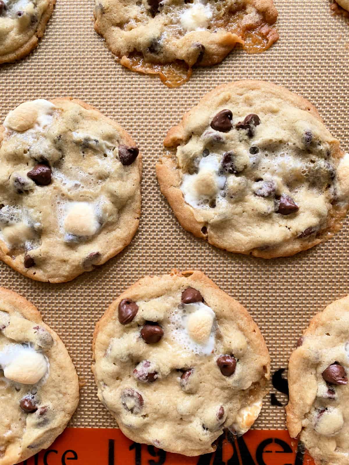 7 baked chocolate chip cookies with marshmallowss on a tan and orange silpat mat.