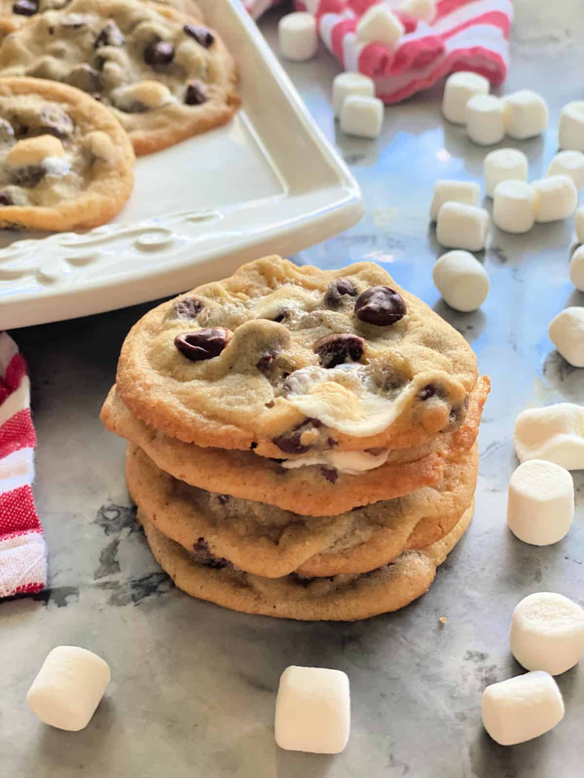 Four stacked chocolate chip marshmallow cookies on a marble counter with mini marshmallows all around.
