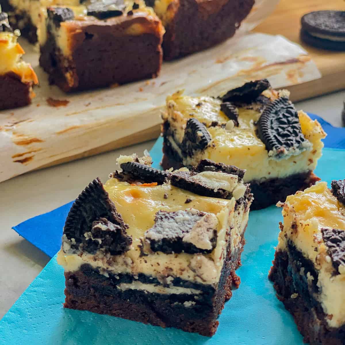 Three slices of brownie topped cheesecake bars with OREOs on a blue napkin.