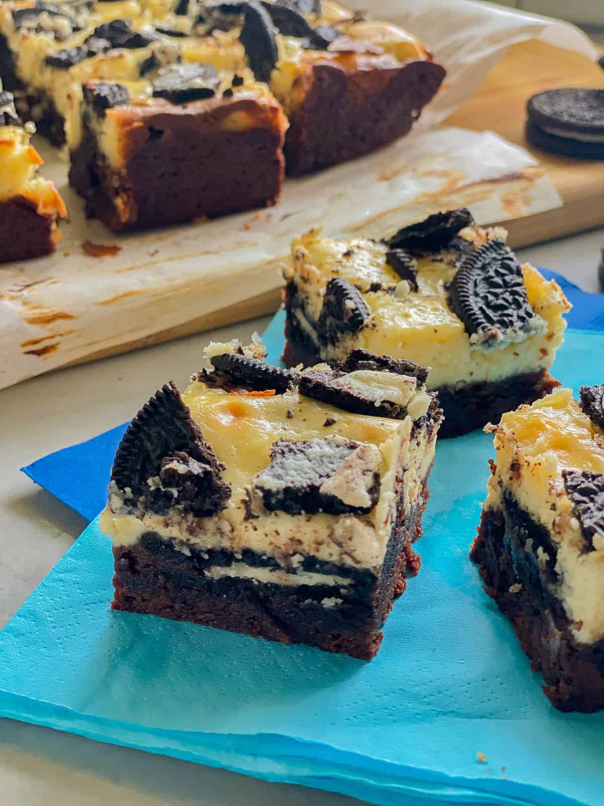 Three slices of brownie topped cheesecake bars with OREOs on a blue napkin.