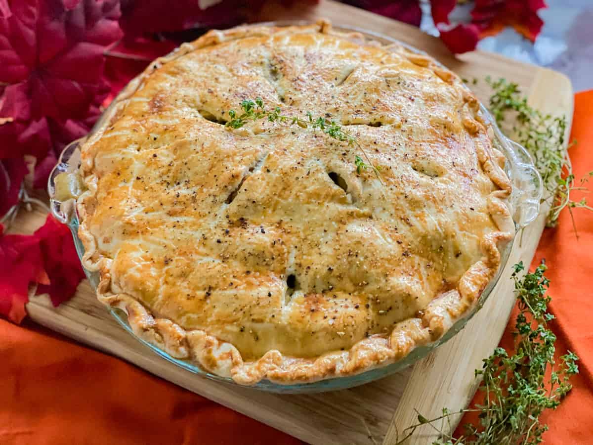 Golden baked pie in a glass dish resting on a wood cutting board with thyme leaves on top.