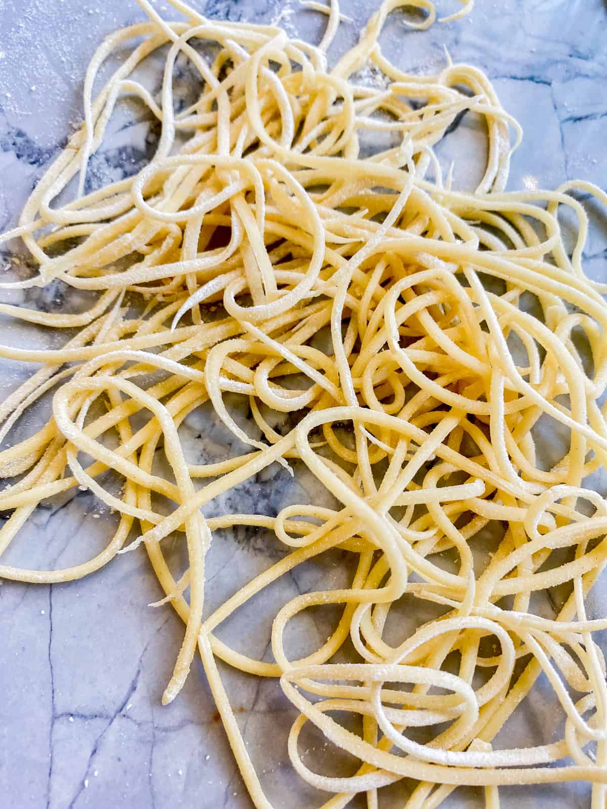 Fresh spaghetti dough tossed with flour on a marble countertop.