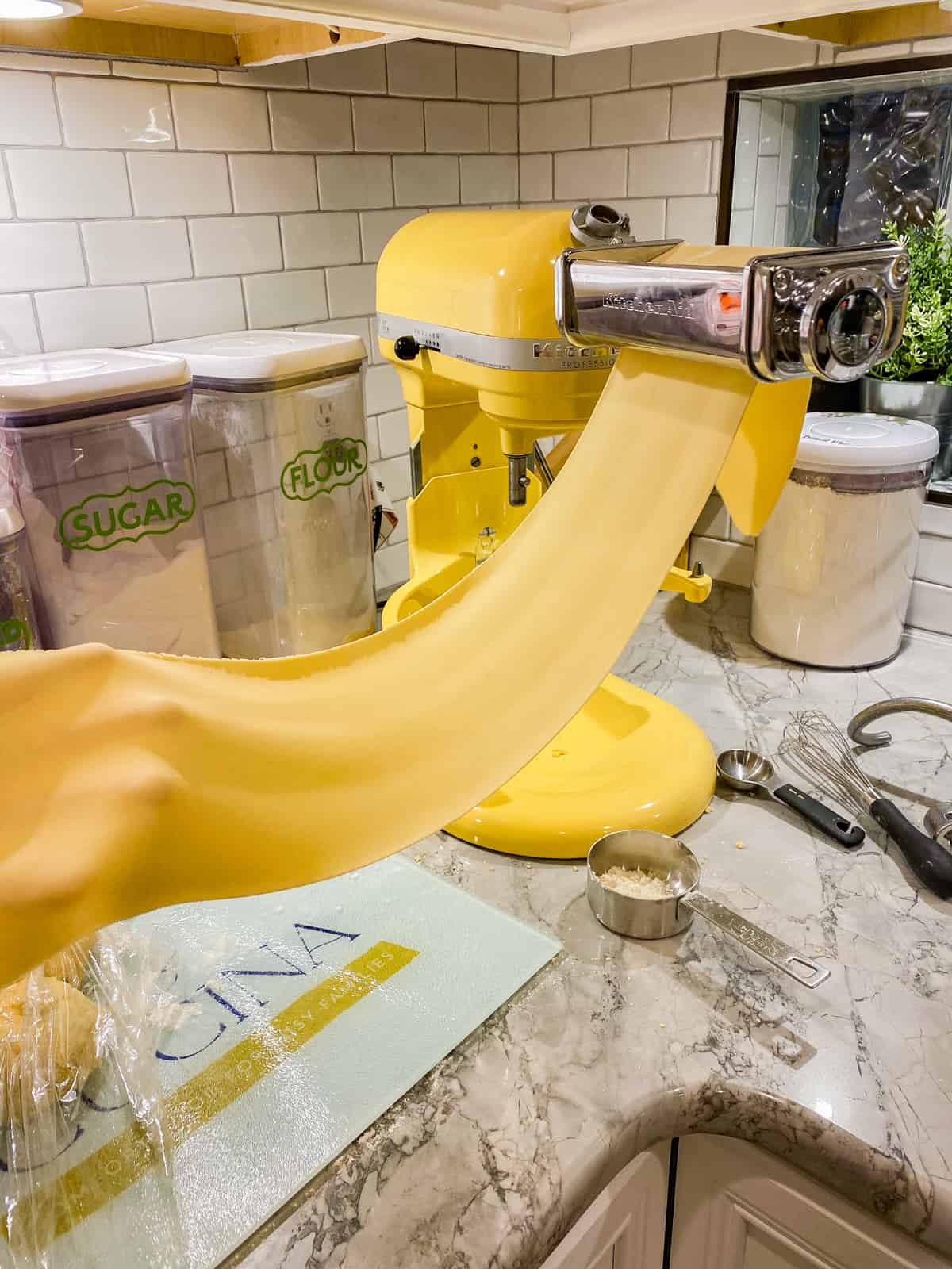 Yellow KitchenAid stand mixer with a hand holding a long sheet of pasta going through a roller.