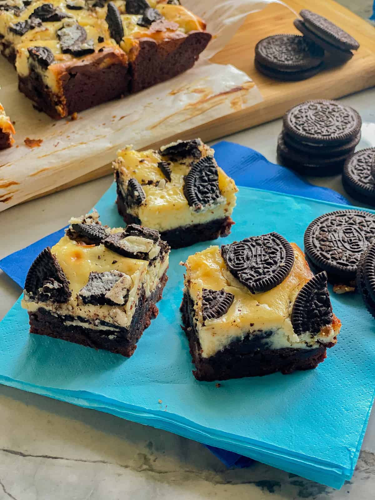 Three slices of brownies topped with cheesecakes and oreos on a bright blue napkin.