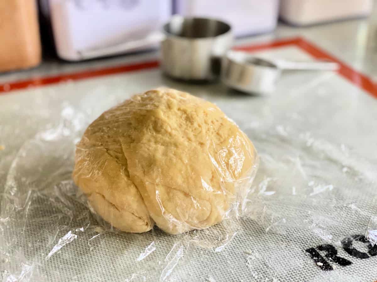 Dough ball wrapped in plastic wrap resting on a silicone baking mat with measuring cups in the background.