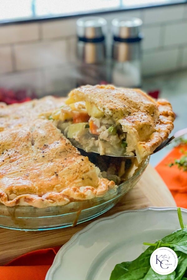 Spatula removing a slice of turkey pot pie from a glass pie plate with a logo on the bottom right corner.