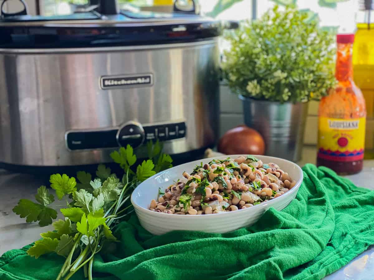 White oval bowl filled with cooked black eyed peas wrapped around green cloth, parsley, and slow cooker in background.