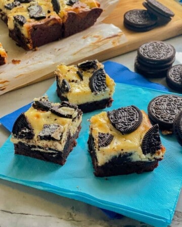 Three slices of brownies topped with cheesecakes and oreos on a bright blue napkin.