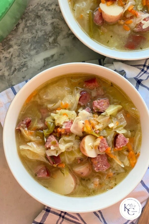 Two white bowls filled with sausage, potatoes, and cabbage with broth with logo on right corner.