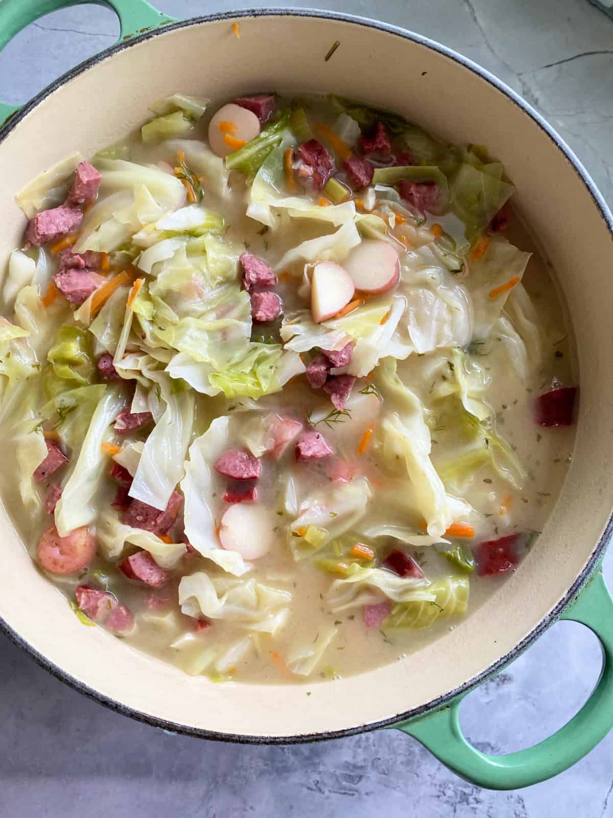 Large green pot filled with cabbage, sausage, potatoes, and broth on a marble countertop.