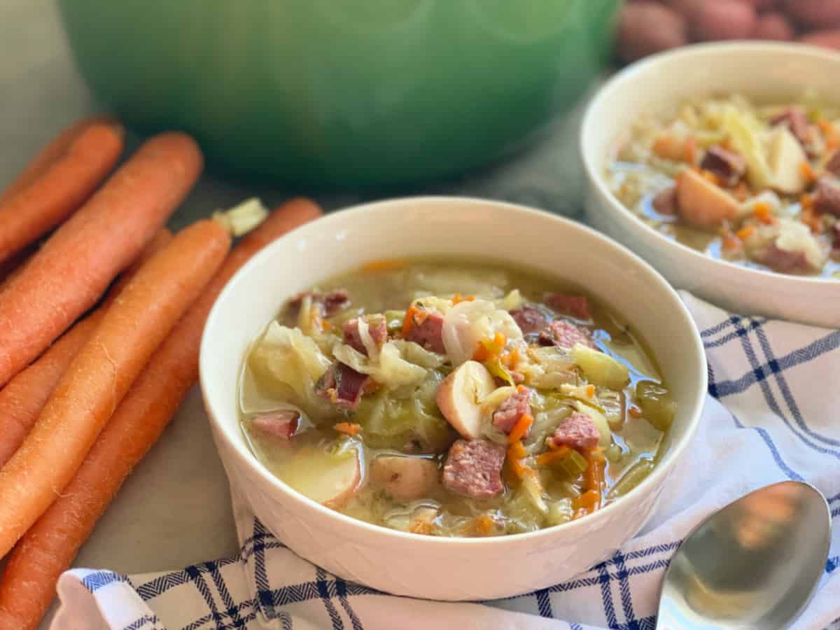 Two white bowls filled with soup next to a pile of carrots and a green pot in the background.