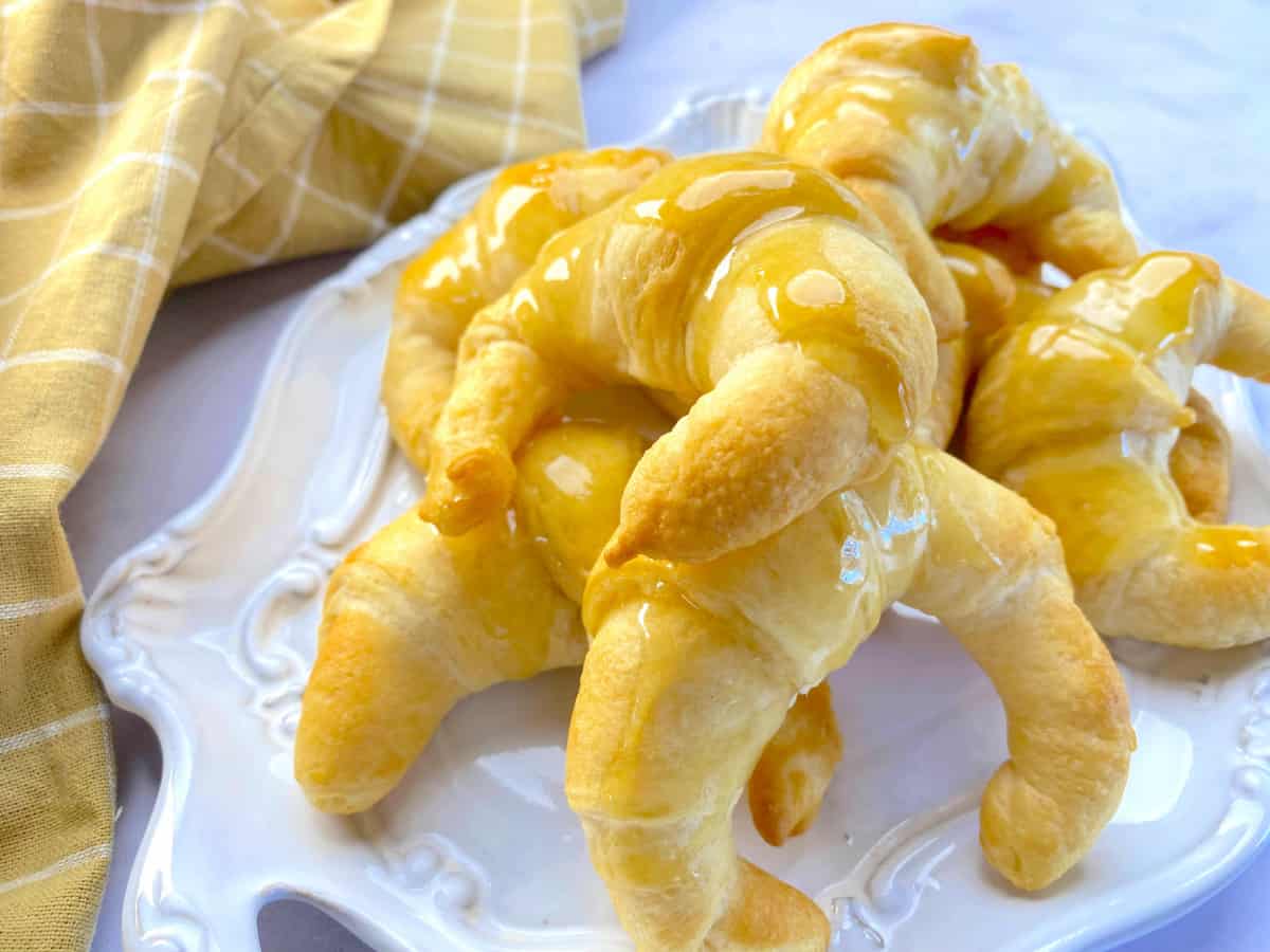 Baked croissants with glaze on a white platter with yello cloth in the background.